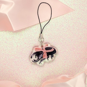 May include: A clear charm with a black stag beetle wearing a pink bow. The charm is attached to a black string.