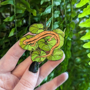Puede incluir: Pin de esmalte dorado con una salamandra sobre un lecho de hojas verdes. La salamandra es naranja y negra con un diseño detallado.