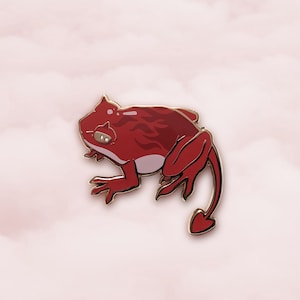 May include: Red and gold enamel pin of a frog with a tail, the frog has a flame-like pattern on its back.