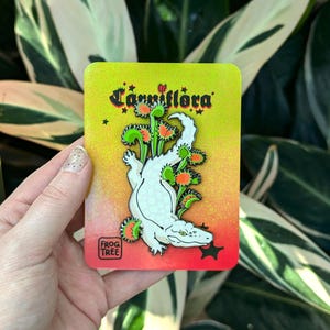 May include: A rectangular pin featuring a white alligator-like creature and Venus flytraps, set against a yellow to red gradient. The pin has the text "Carniflora" and the "Frog Tree" logo.