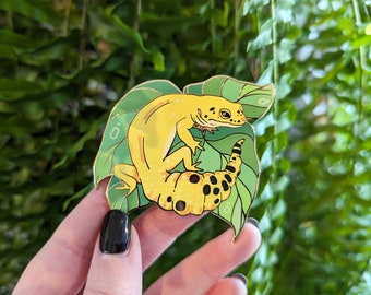 May include: A gold-coloured enamel pin depicting a yellow and black gecko on a green leaf. The gecko is curled up on the leaf, with its tail wrapped around its body.