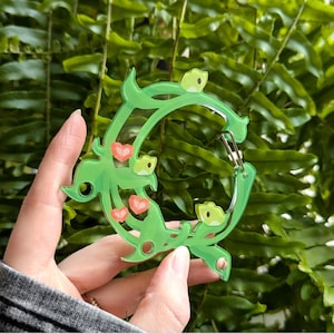 Puede incluir: Un mosquetón verde acrílico con un diseño floral, con pequeños corazones rojos y detalles en verde claro. El mosquetón tiene un cierre de metal plateado y se sostiene contra un fondo de follaje verde.