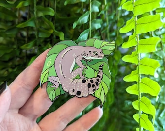 May include: A pink and green enamel pin featuring a gecko on a leaf. The gecko has black spots and a curled tail.