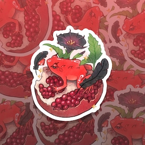 Op de afbeelding: Een sticker met een rode kikker die in een granaatappel zit. De kikker heeft een geïrriteerde uitdrukking en wordt omringd door zwarte veren, groene bladeren en een donkerpaarse bloem. De sticker heeft een witte rand.