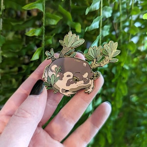 Pode incluir: Um broche de esmalte dourado com um desenho de sapo de desenho animado com marcas marrons e brancas, sentado em um leito de folhas verdes. O sapo tem uma expressão rabugenta e usa uma pequena coroa.