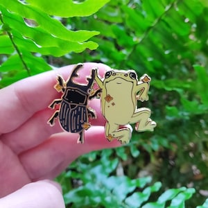 May include: Two enamel pins, one of a black beetle and one of a yellow frog, both with gold accents. The pins are on a hand against a green leafy background.