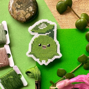 Könnte beinhalten: Ein grünes Kreuzstich-Design eines Cartoon-Charakters mit weißem Rand. Das Bild zeigt auch Spulen mit grünem und braunem Garn, einen polierten Stein und rosa Blüten. Der Hintergrund ist grün.