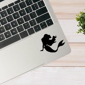 May include: A silver laptop computer with a black keyboard and a black silhouette of a mermaid on the trackpad.