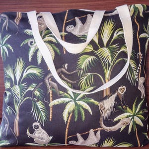 Peut inclure: Un sac cabas noir avec des anses blanches présente un imprimé tropical de palmiers, de singes et de paresseux. L'imprimé est dans des tons de vert, de brun et de blanc.