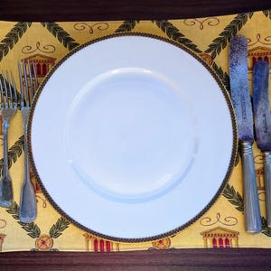 Op de afbeelding: Een wit dinerbord met een gouden rand ligt op een gele placemat met een herhalend patroon van groene bladeren en gouden gebouwen. Twee zilveren vorken en twee zilveren messen liggen rechts van het bord.