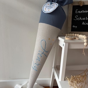 Könnte beinhalten: Eine Schultüte aus Stoff mit einem blauen Musselin-Oberteil und einem beigen Waffelstoff-Unterteil. Der Name Henry ist in blauer Schreibschrift gestickt, ergänzt durch goldene Sterne. Ein rundes weißes Schild trägt die Aufschrift Schulkind 2026.