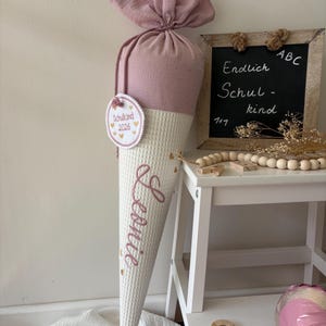 Könnte beinhalten: Eine kegelförmige Geschenktüte aus Stoff mit einem altrosa Oberteil und einem weißen Unterteil in Waffelstruktur. Die Tüte ist mit dem Namen Leonie in rosa Schreibschrift personalisiert und hat einen runden Anhänger mit Schulkind 2026.