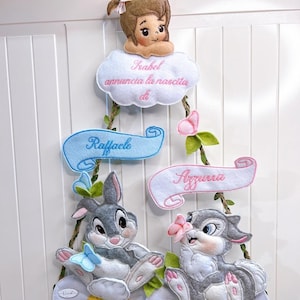 Op de afbeelding: Een vilten baby-aankondigingsdecoratie met een meisje op een wolk, banners met namen en twee cartoonkonijnen op madeliefjes. De tekst op de wolk luidt "Isabel kondigt de geboorte van" aan. De banners tonen de namen "Raffaele" en "Azzurra".