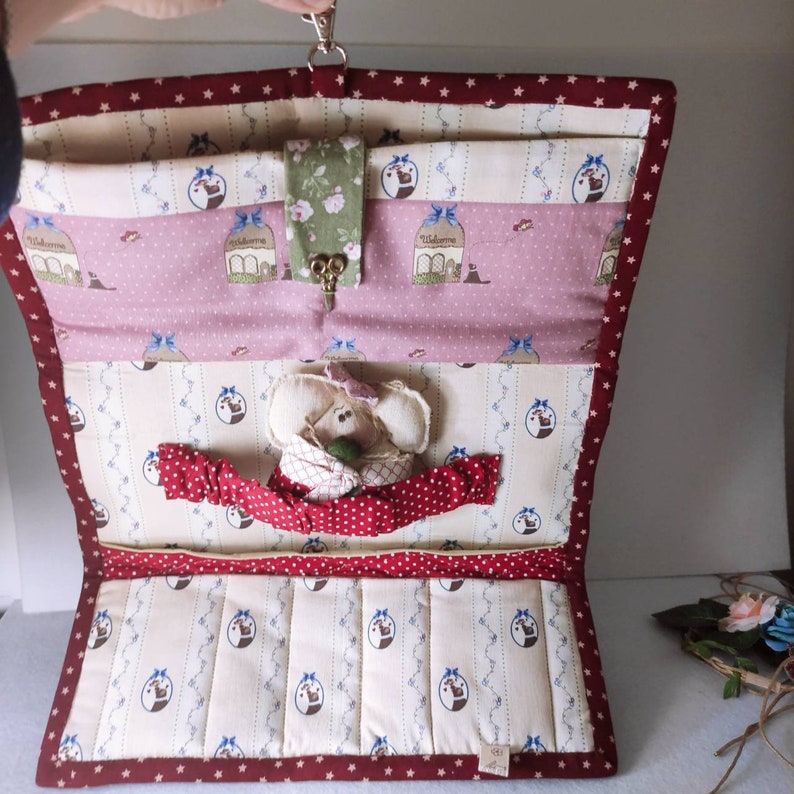 May include: A red and white quilted fabric organizer with a small stuffed animal inside. The organizer has a zipper closure and a keyhole on the front. The organizer is decorated with a variety of patterns, including stars, stripes, and polka dots.