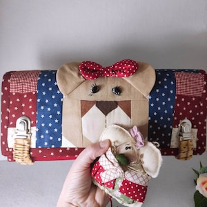 May include: A red, white, and blue patchwork fabric pouch with a bear face on the front. The bear has a red bow and black eyes. A small, white, patchwork fabric doll is in the hand of a person holding the pouch.