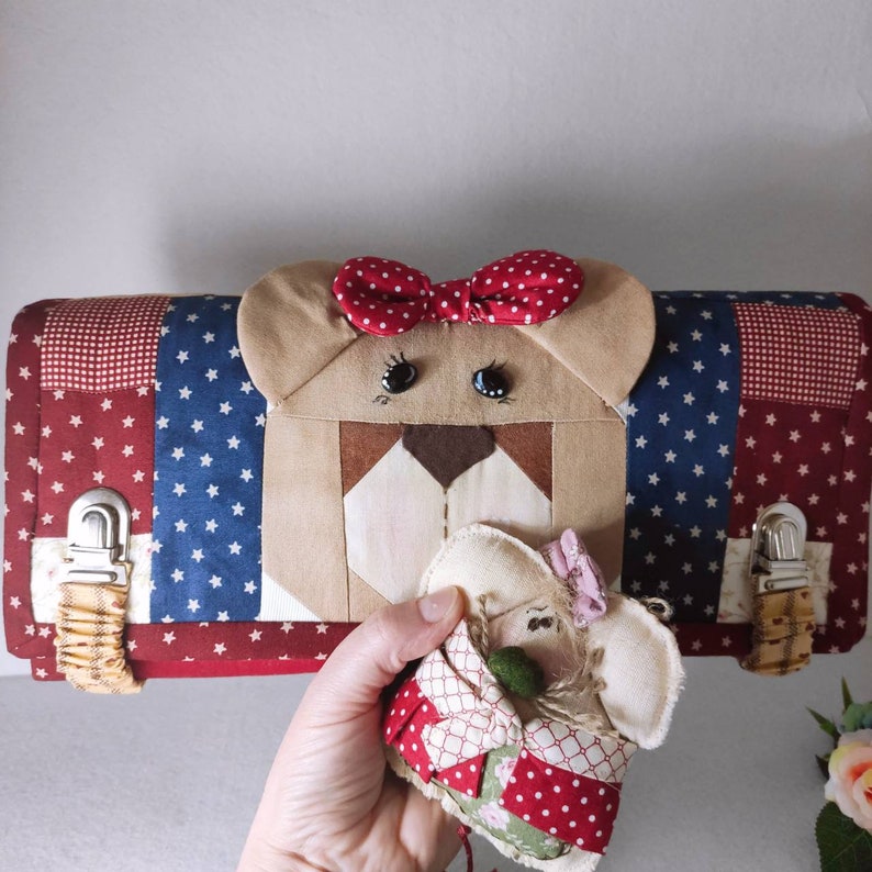 May include: A red, white, and blue patchwork fabric pouch with a bear face on the front. The bear has a red bow and black eyes. A small, white, patchwork fabric doll is in the hand of a person holding the pouch.