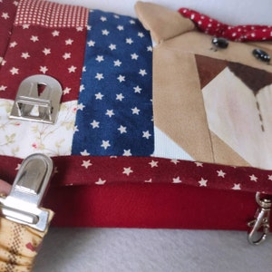 May include: A red, white, and blue patchwork fabric bag with a brown dog face on the front. The bag has a zipper closure and a metal clasp. The bag is decorated with white stars and red and white checkered fabric.