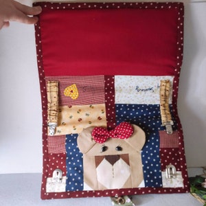 May include: A red, blue, and yellow patchwork baby activity quilt with a brown bear face in the center. The quilt has a variety of textures and patterns, including stars, polka dots, and flowers. The bear has a red bow and brown eyes. The quilt has two yellow straps with clips on each side.