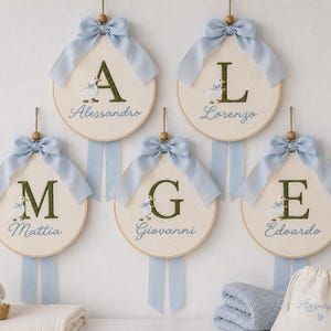 Può includere: Cinque telai da ricamo circolari con tessuto color crema, ciascuno ricamato con una grande lettera verde scuro, una piccola anatra bianca e un nome in corsivo blu. Ogni telaio è sormontato da un fiocco in nastro di raso azzurro e una perla.