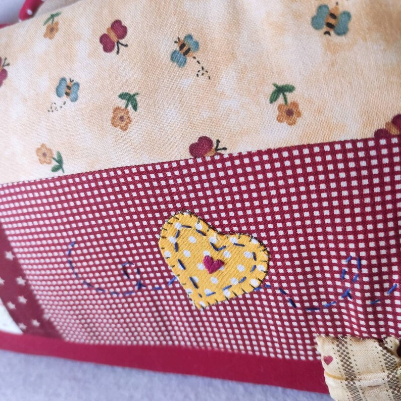 May include: A red and white checkered fabric with a yellow heart with white polka dots and a red heart in the center. The heart is stitched with blue thread. The fabric has a white star in the corner.