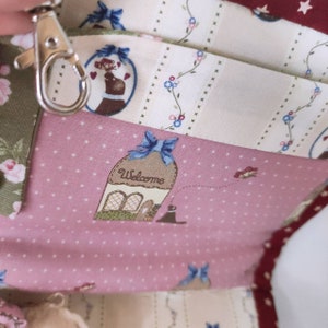 May include: A pink fabric bag with a patchwork design featuring a house with a welcome sign, a blue bow, and a small bird. The bag has a key chain attached to it.