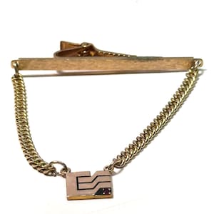 May include: A gold-tone tie bar featuring a textured horizontal bar with a spring-loaded clip on the back. A gold-tone curb chain connects the bar to a rectangular charm with black geometric line detailing and a small embedded red gemstone.