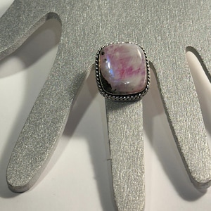 May include: A square-faced ring with a silver band and a large, light pink and white stone. The stone is set in a silver frame with a twisted rope design. The ring is displayed on a silver hand-shaped display.