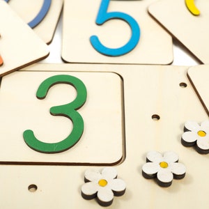 May include: Wooden number tiles with colorful numbers, including 1, 2, 3, 4, and 5. The tiles are arranged in a random order on a white surface. There are also small white flowers with yellow centers on the surface.