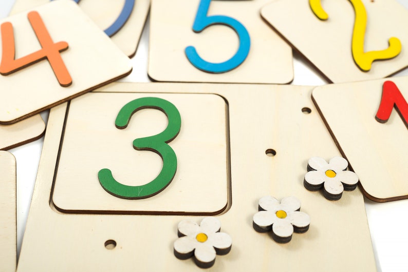 May include: Wooden number tiles with colorful numbers, including 1, 2, 3, 4, and 5. The tiles are arranged in a random order on a white surface. There are also small white flowers with yellow centers on the surface.