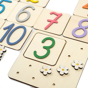May include: Wooden number tiles with colorful numbers from 1 to 10. The tiles are arranged in a grid pattern. There are also small white flower-shaped pieces.