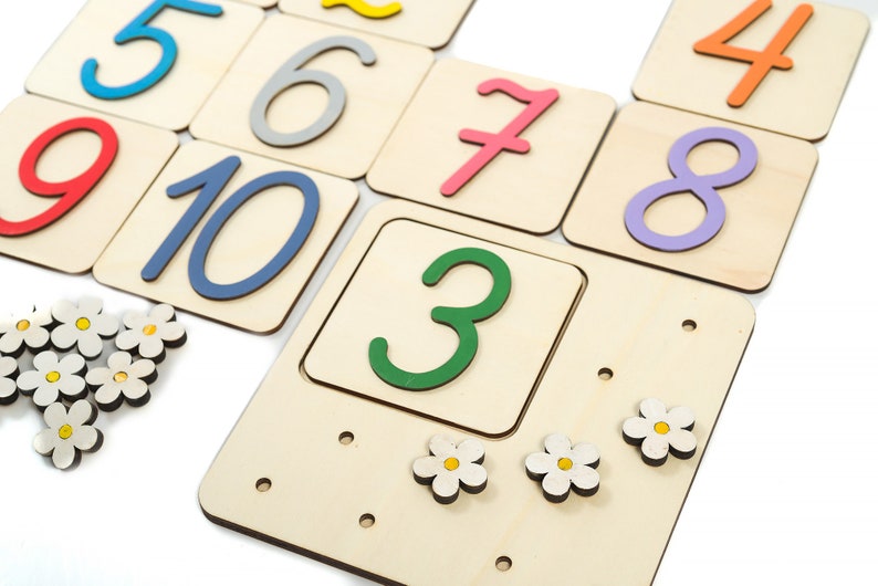 May include: Wooden number tiles with colorful numbers from 1 to 10. The tiles are arranged in a grid pattern. There are also small white flower-shaped pieces.