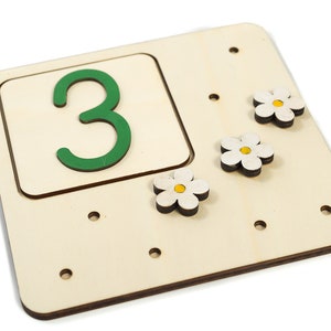 May include: Wooden board with a green number 3 in a cutout square. There are three white flower shapes with yellow centers attached to the board.