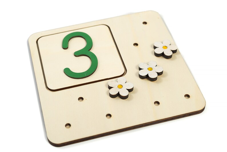 May include: Wooden board with a green number 3 in a cutout square. There are three white flower shapes with yellow centers attached to the board.