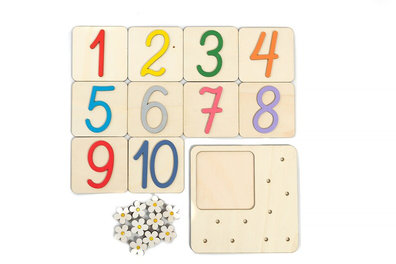 May include: Wooden number tiles with colorful numbers 1 through 10. The tiles are arranged in three rows. There is a wooden frame with a blank space and holes around the edge. There are also small white wooden flowers with yellow centers.