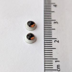 May include: Two brown and black plastic safety eyes with white plastic rims, measuring approximately 1 cm in diameter, on a white surface.