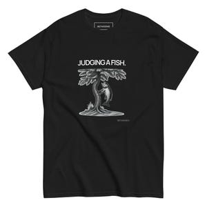 以下が含まれることがあります： 木にぶら下がっている魚のグラフィックが特徴の黒いTシャツ。画像の上には「JUDGING A FISH.」というテキストがあります。ブランド名「BETWEENIE」はシャツの上部と下部に印刷されています。