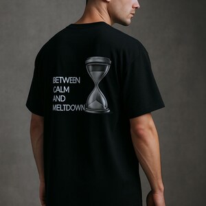 以下が含まれることがあります： 「BETWEEN CALM AND MELTDOWN」の文字と砂時計のグラフィックが入った黒いTシャツ。柔らかい素材でできており、クルーネックのデザインです。画像はTシャツの背面を示しています。