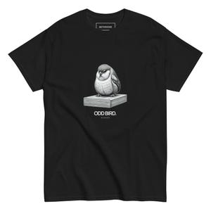 以下が含まれることがあります： 黒いTシャツ。本の上に止まっているふっくらとした鳥の精巧なイラストが描かれています。「ODD BIRD」の文字が下にプリントされています。Tシャツはクルーネックで半袖です。