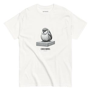 以下が含まれることがあります： 白いTシャツ。本の上に止まっている丸い鳥の白黒イラスト。鳥の下には「ODD BIRD」の文字がプリントされています。Tシャツは半袖で、クルーネックです。