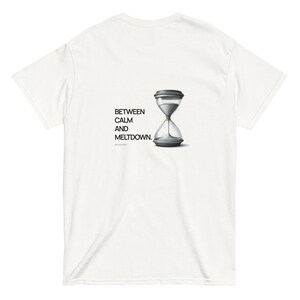 以下が含まれることがあります： 白地のTシャツ。黒と白の砂時計のグラフィックと「BETWEEN CALM AND MELTDOWN」の文字がプリントされています。砂時計は詳細に描かれており、上部と下部は暗い色です。このTシャツは柔らかい素材で作られています。