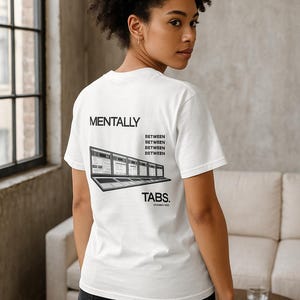以下が含まれることがあります： 「MENTALLY BETWEEN BETWEEN BETWEEN TABS」の文字が入った白いTシャツ。テキストの下には、複数のタブが開いているラップトップのグラフィックが印刷されています。Tシャツは人が着ています。