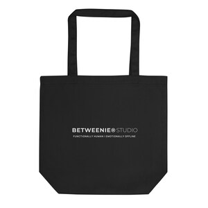 以下が含まれることがあります： 長方形のボディと2つのハンドルが付いた黒いトートバッグ。「BETWEENIE® STUDIO」の白い文字と「FUNCTIONALLY HUMAN | EMOTIONALLY OFFLINE」のスローガンが特徴です。