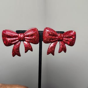 May include: A pair of red glitter bow earrings. The earrings are shaped like bows with a textured surface and are mounted on black earring stands. The bows have a shiny, sparkly appearance.