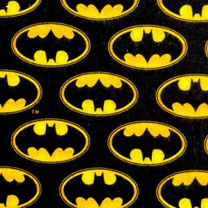 Puede incluir: Tela negra con un patrón repetido del logotipo de Batman en amarillo. El logotipo tiene forma ovalada con una silueta de murciélago en el centro. El símbolo de la marca TM es visible en algunos logotipos.