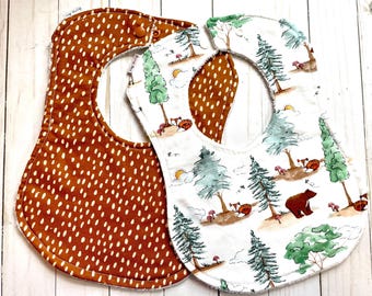 May include: Two baby bibs. One is brown with white polka dots, and the other is white with a forest scene featuring trees, a bear, foxes, and mushrooms. Both bibs have a snap closure.