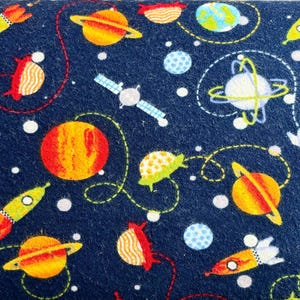 May include: A dark blue fabric featuring a repeating space pattern with yellow and orange rockets, various planets with rings, a satellite, and small white dots representing stars. The design includes dashed orbital lines connecting the celestial objects.
