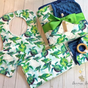 May include: A baby gift set featuring a bib, burp cloth, blanket, and teether. The items are decorated with a pattern of green and blue sea turtles on a white background. The blanket is tied with a green ribbon.