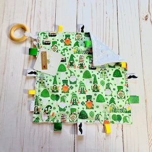 Könnte beinhalten: Ein grünes und weißes Babyspielzeug mit einem Holzring, einem weißen Etikett mit der Aufschrift "Little Bear" und verschiedenen grünen, gelben und weißen Bändern. Das Spielzeug ist mit einem Muster aus Gnomen verziert, die grüne Hüte tragen und Kleeblätter halten. Das Spielzeug hat eine weiße, weiche, strukturierte Seite.