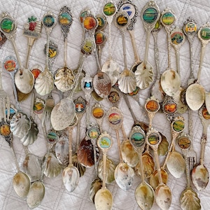 Puede incluir: Una colección de cucharas de recuerdo vintage con mangos decorativos y escudos de esmalte de colores. Las cucharas presentan varios diseños, incluyendo monumentos, paisajes y emblemas. Algunas cucharas tienen cuencos en forma de concha.