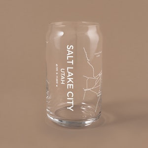 May include: A clear glass can with a white outline of the Salt Lake City, Utah map. The text "SALT LAKE CITY" is printed vertically on the left side of the can. The text "UTAH" is printed below the city name. The coordinates "40.7595 N, 111.8937 W" are printed below the state name. The text "Great Salt Lake" is printed at the top of the map.
